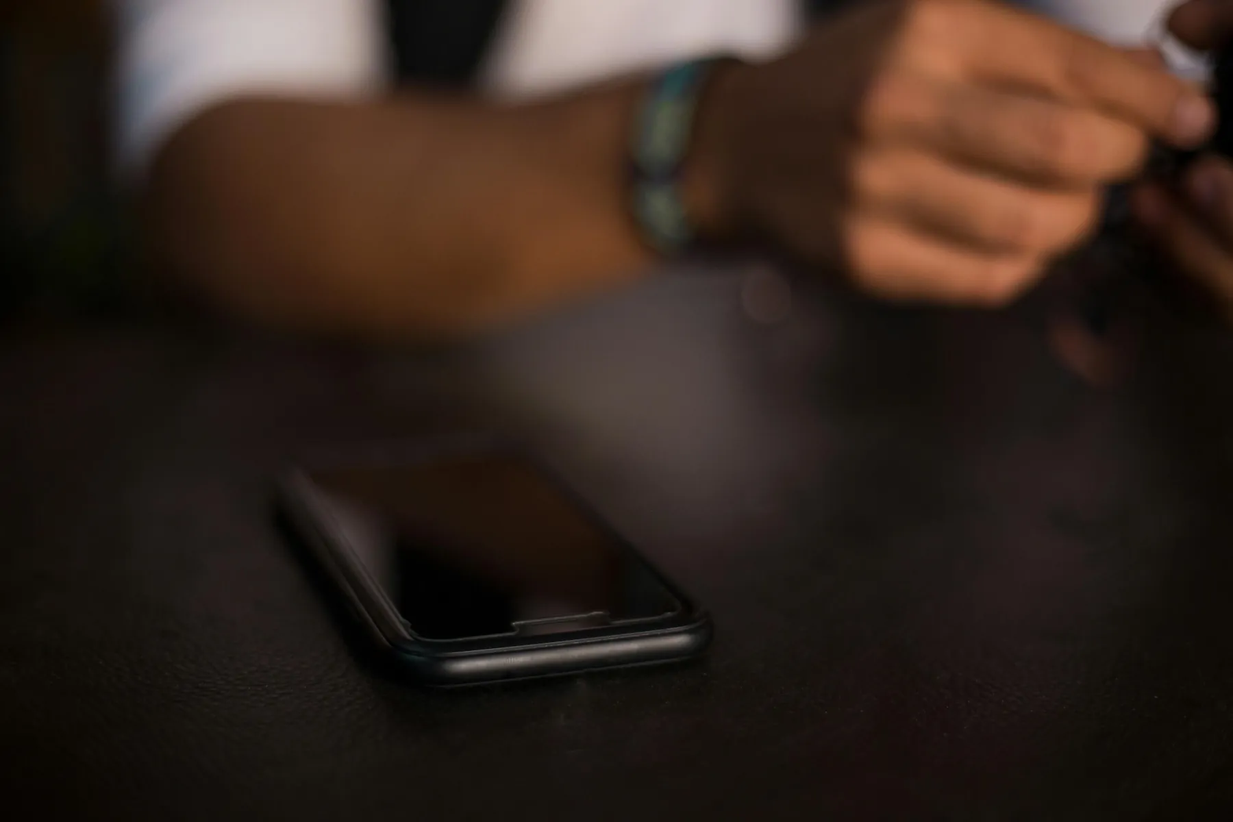 Free A smartphone rests on a table with a person holding an object in the blurred background, in a dark indoor setting.